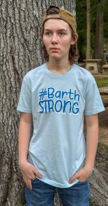A teenage boy wearing a “#Barth Strong” shirt stands outdoors with his hands in his pockets.