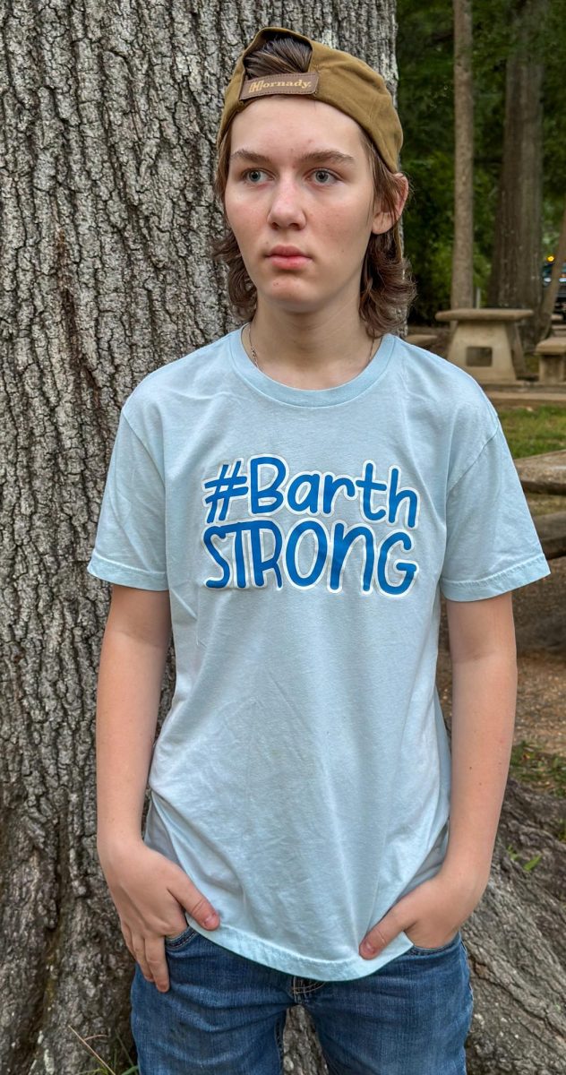 A teenage boy wearing a “#Barth Strong” shirt stands outdoors with his hands in his pockets.