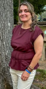 A woman in a burgundy blouse stands near a tree, smiling slightly.