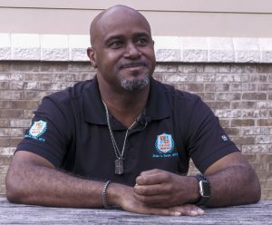 A man wearing a black polo sits at a picnic table in front of a brick wall.