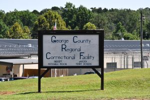Sign outside the George County Regional Correctional Facility with fencing and buildings visible in the background.