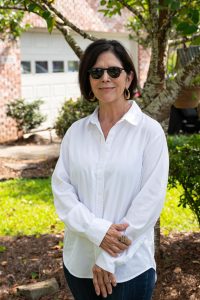 A woman wearing a white shirt and sunglasses, standing outside near her home.