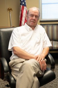 A man in a white shirt speaking while sitting in a chair in an office.