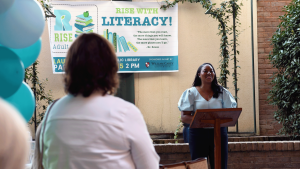 A woman with long dark hair wearing a light blue blouse stands behind a wooden podium, smiling as she addresses an audience. A banner behind her reads “Rise with Literacy!” with illustrations of stacked books and a quote from Dr. Seuss. Blue and white balloons frame the scene, and the audience is partially visible in the foreground. The event appears to take place outdoors in a courtyard surrounded by brick walls and green plants.