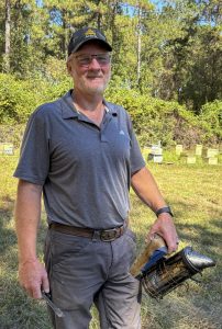A man in a dark gray hat and glasses holds a bee smoker while standing near hives in a grassy clearing.