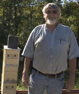 A bearded man wearing glasses and a gray shirt stands near stacked wooden bee boxes in a sunny field.
