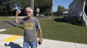An older man with short gray hair stands on a sidewalk beside a residential street. He wears glasses, a gray T-shirt with yellow lettering that reads “SPRINGSTEEN STREET BAND,” and jeans. He gestures behind him with his thumb, as if pointing toward a location in the neighborhood under bright sunlight.
