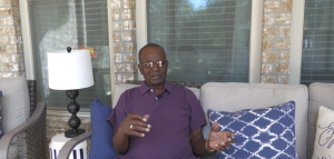 An older man wearing glasses and a purple polo shirt sits on a beige outdoor sofa on a brick porch. He gestures with his hands while speaking, framed by decorative pillows and a lamp beside him. The background shows large windows with blinds partially drawn, reflecting daylight.