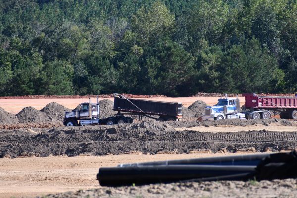 Two large dump trucks and several mounds of brown dirt sit on a cleared construction site surrounded by a dense tree line. The trucks appear mid-operation under a bright blue sky, with tire tracks and layers of freshly moved earth visible across the foreground.