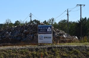 A roadside construction area with piles of gray rocks stacked behind a blue-and-white sign reading “Pearl River County Industrial Park.” Wooden utility poles and overhead wires frame the scene against a clear blue sky and a backdrop of trees.