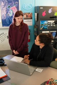 A teenage girl in a black hoodie interacts with another student in a maroon sweater at a desk.