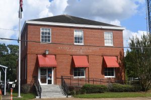 A two-story red brick building with red awnings and white trim houses the former Poplarville Post Office. An American flag flies in front of the building under a partly cloudy sky.