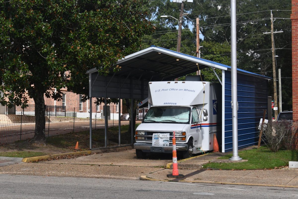 A white postal van labeled “U.S. Post Office on Wheels” is parked under a blue metal awning beside a brick building, serving as a temporary post office.