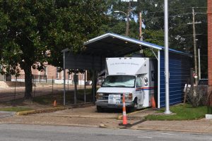 A white postal van labeled “U.S. Post Office on Wheels” is parked under a blue metal awning beside a brick building, serving as a temporary post office.
