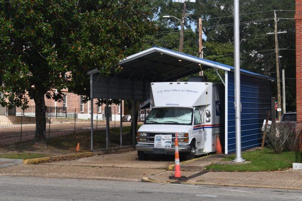A white postal van labeled “U.S. Post Office on Wheels” is parked under a blue metal awning beside a brick building, serving as a temporary post office.
