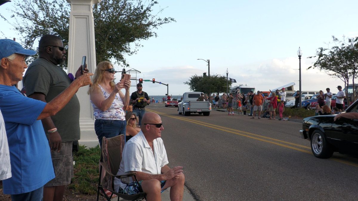 A group of spectators sits on a seawall under tents and palm trees, watching classic cars cruise by during the annual Cruisin’ the Coast celebration in Long Beach.