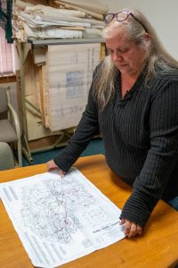 A woman with long gray hair examines a large map laid out on a wooden table.