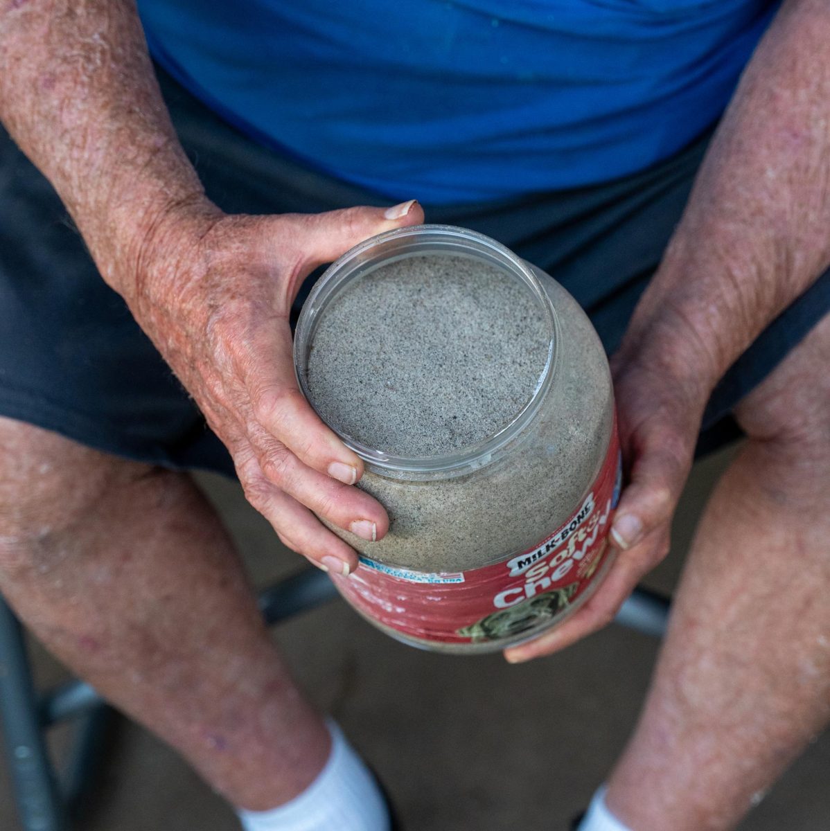 Close-up for a man in a blue shirt and shorts holding a plastic jar full of fine sand.