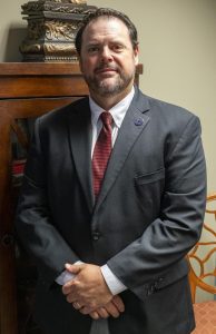 Man with short brown hair and beard wearing a dark suit and red tie, standing indoors with hands clasped.