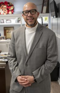 A bald man in glasses and a gray suit standing in an office with hands clasped.