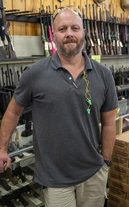 A man wearing a gray polo shirt stands in front of a gun counter lined with rifles.
