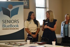 Two women stand beside a sign that reads “Seniors BlueBook: Resources for Aging Well.” A table in front of them holds pamphlets and drinks.