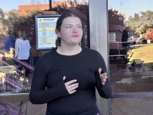 A woman in a black long-sleeve shirt stands in front of a gym window with a facility hours sign behind her while she gestures with her hands.