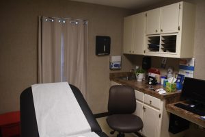 Medical exam room with exam table, rolling chair, cabinets, countertop with supplies, and a computer workstation.