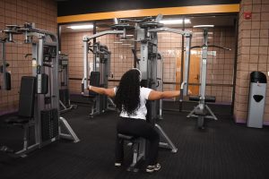 A woman sits on a chest fly machine inside a gym while holding the handles at shoulder height during a workout.
