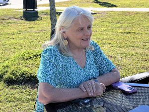 A woman with white hair in a green patterned blouse speaks while seated outdoors at a black metal table with a phone and glasses in front of her.