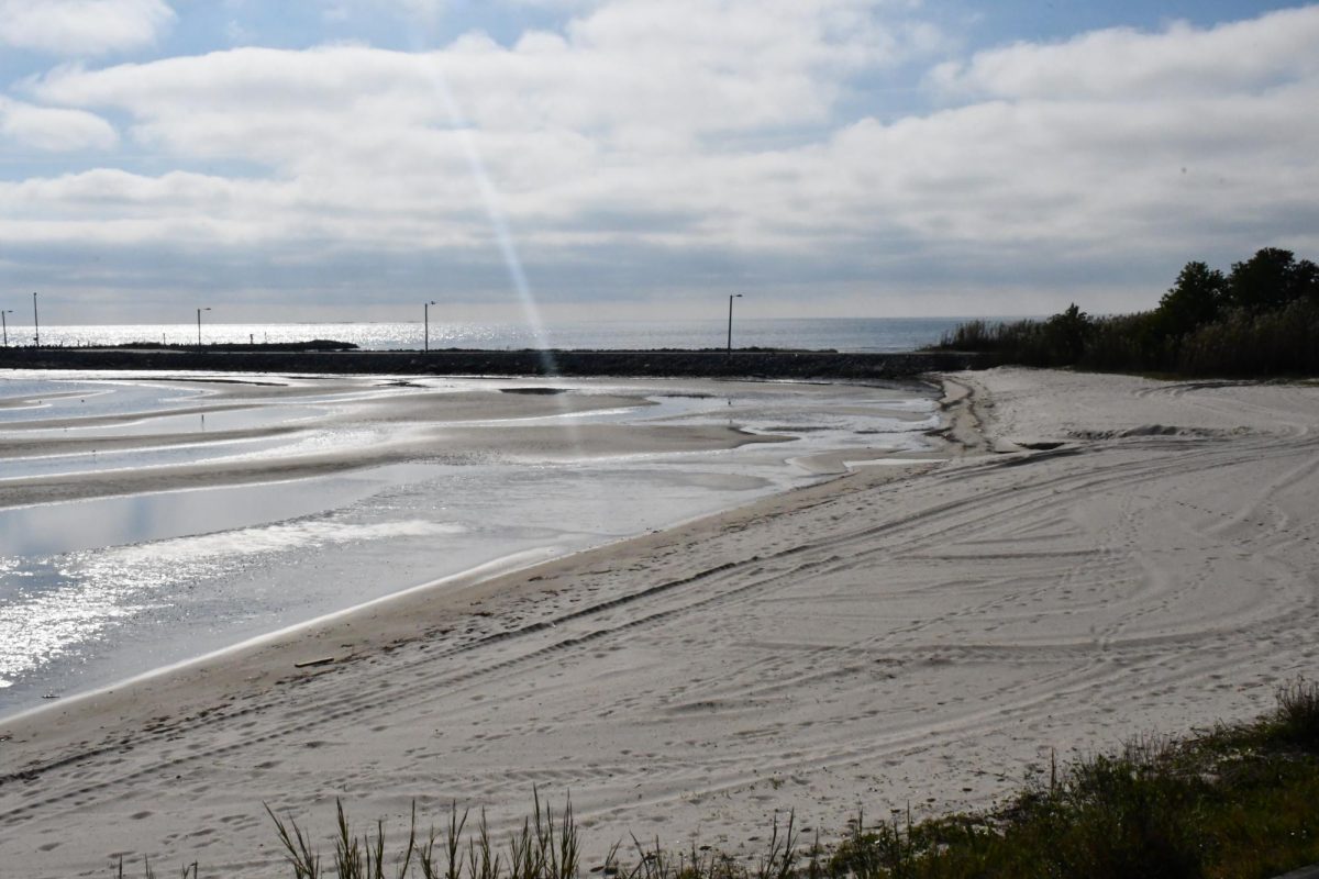 A sandy beach with visible water ripples and tide pools under a partly cloudy sky, with a rock jetty and a few light posts along the shoreline.
