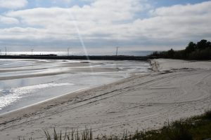 A sandy beach with visible water ripples and tide pools under a partly cloudy sky, with a rock jetty and a few light posts along the shoreline.