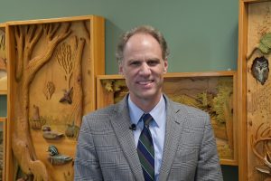 A man wearing a gray plaid suit jacket, blue shirt and striped tie stands indoors in front of carved wooden nature-themed wall art, smiling at the camera.