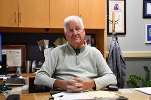 A man with white hair sits at a desk with his hands folded, surrounded by office items including a computer monitor, framed certificates and children’s drawings on the wall behind him.