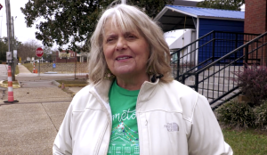 A woman with shoulder-length white hair wearing a white jacket and greet shirt stands on a sidewalk in front on a brick building.