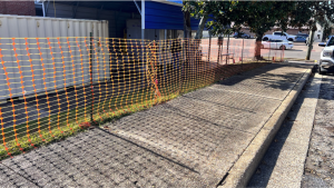 A plastic orange safety fence is hung along a sidewalk in front of a metal overhang.