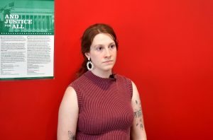Erin Hensley stands against a red wall during an interview. She is wearing a sleeveless maroon top and earrings.