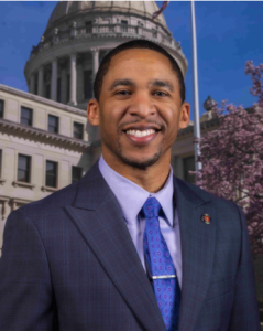 A man wearing a suit and tie poses in front of the Mississippi Capitol.