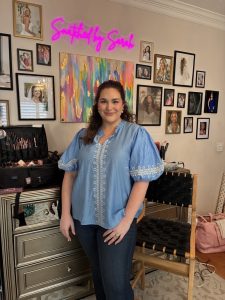 A woman with dark hair and a blue, short-sleeve shirt stands in front desk covered in beauty supplies, surrounded by framed photos and artwork.