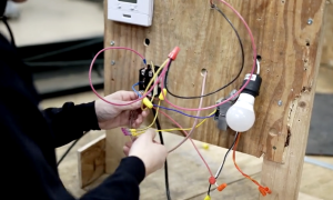 Hands adjust multicolored electrical wires on a training board with a light bulb during HVAC instruction.