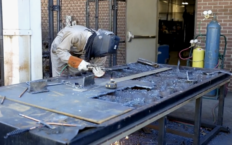A student wearing protective gear welds metal at a workbench during hands-on training.