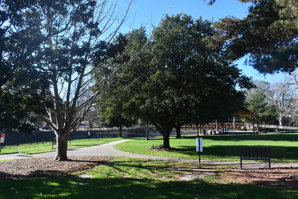 A paved walking path curves through Town Square Park in downtown Hattiesburg, with benches, mature trees and open green space on a sunny day.