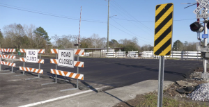 Asphalt extends across a railroad crossing, blocked by road closed signs.
