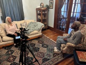 A woman sits on a couch in a living room being interviewed by a reporter with a video camera.