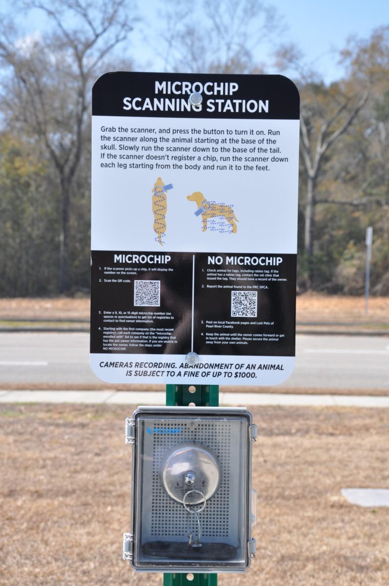 A black-and-white sign titled “Microchip Scanning Station” is mounted on a green post outdoors near a roadway. The sign includes printed instructions, small illustrations of a dog and cat being scanned and two QR codes. A locked metal box containing the scanner is attached below the sign.