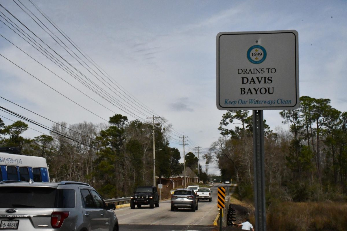Cars drive across a two-lane bridge near a roadside sign reading “Drains to Davis Bayou: Keep Our Waterways Clean.”