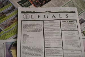 A newspaper, turned to the legal and public notices page, sits on a table covered with other pages of the same newspaper.