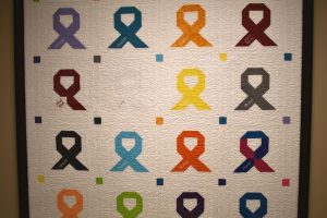 A framed quilt with colorful cancer awareness ribbons and labels for different cancer types.