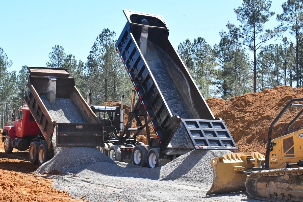 Dump trucks unload gravel at a construction site surrounded by pine trees.