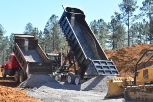 Dump trucks unload gravel at a construction site surrounded by pine trees.